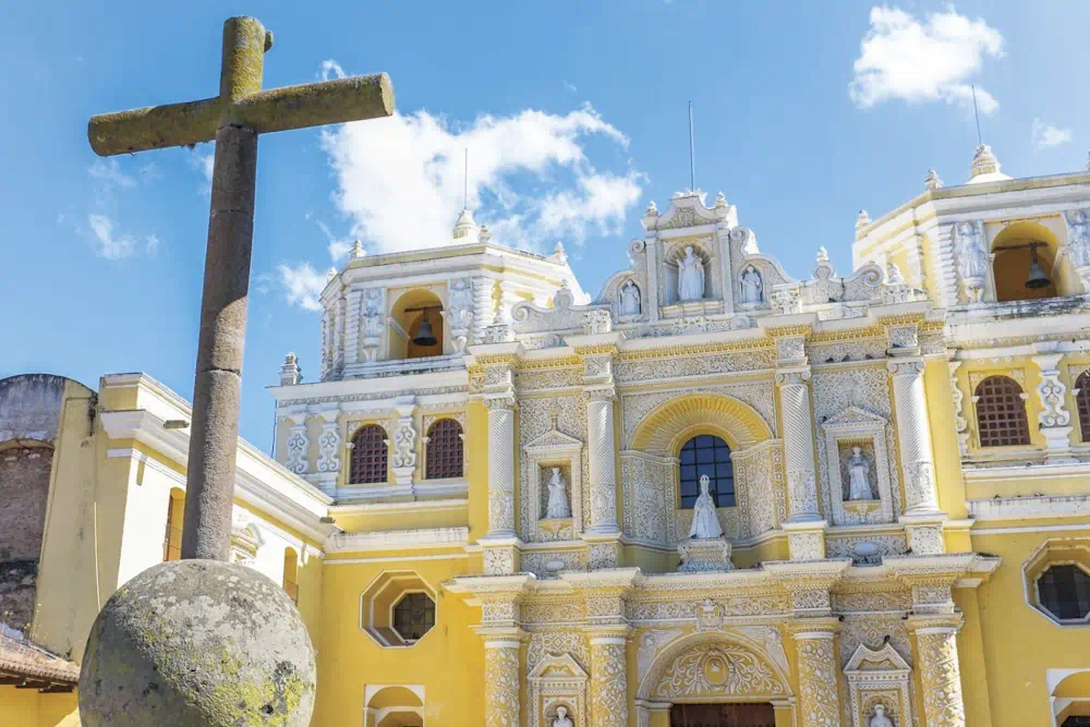Stone cross before ornate yellow colonial church